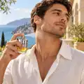 Uomo con camicia di lino che spruzza Azzaro Wanted Man 50 ml sul collo su una terrazza affacciata sul mare
